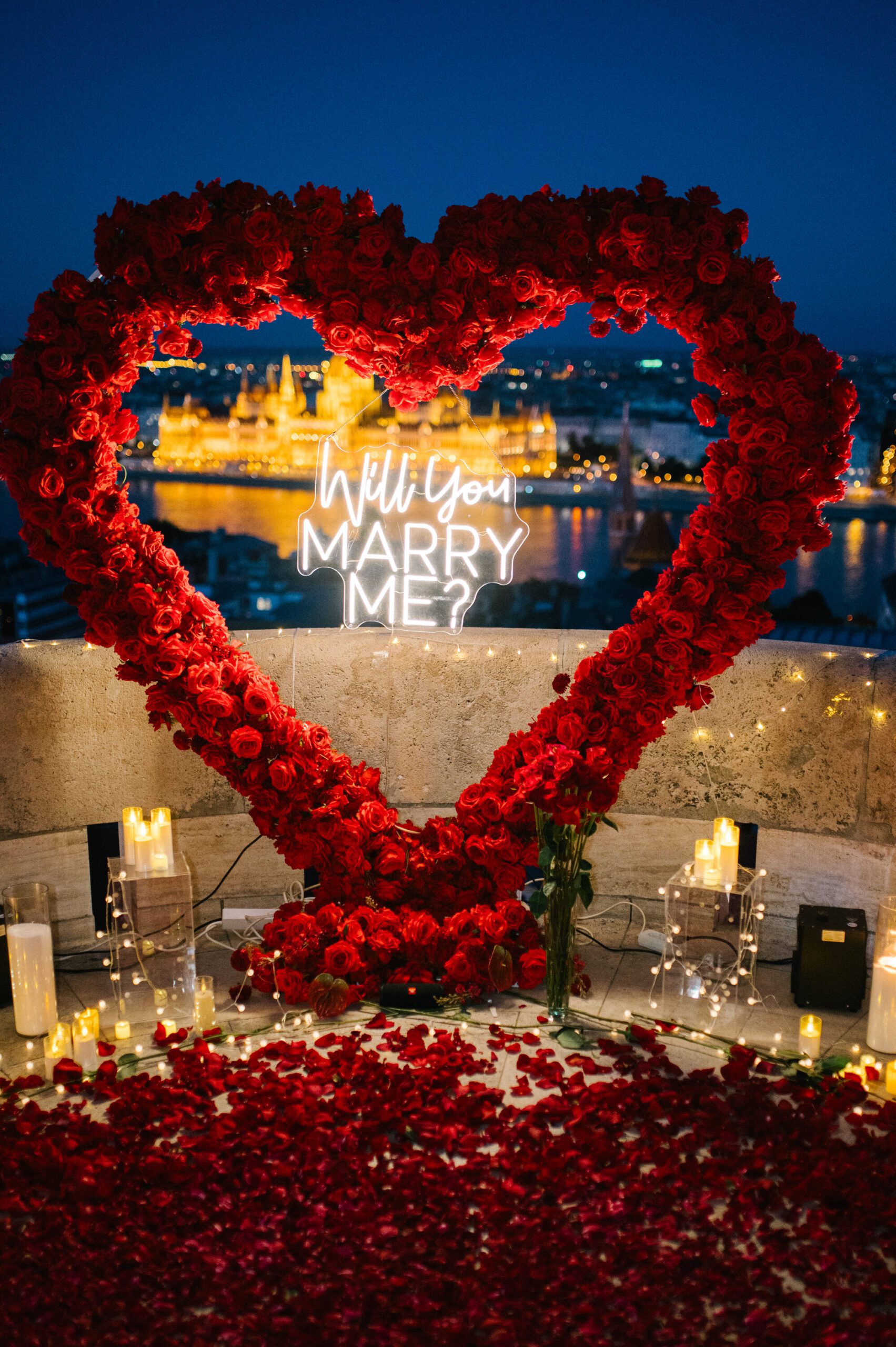Luxury marriage proposal setup with a red rose heart, candles and “Will You Marry Me?” sign overlooking the Liberty Bridge in Budapest