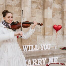 Female violinist playing romantic music during a surprise marriage proposal in Budapest with red heart balloons and glowing 
